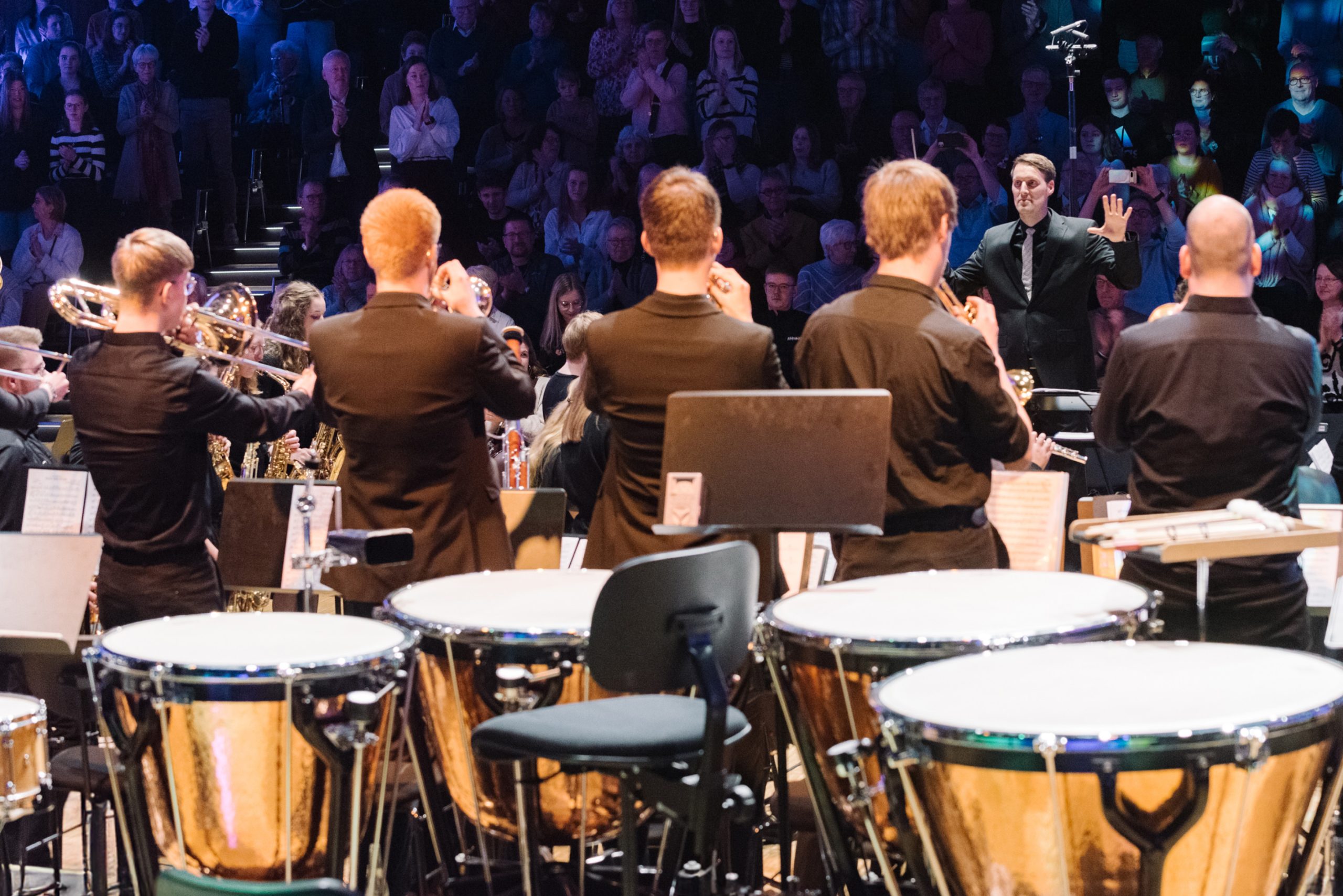 Ein Dirigent steht vor einem Orchester. Im Hintergrund ist das Publikum zu sehen